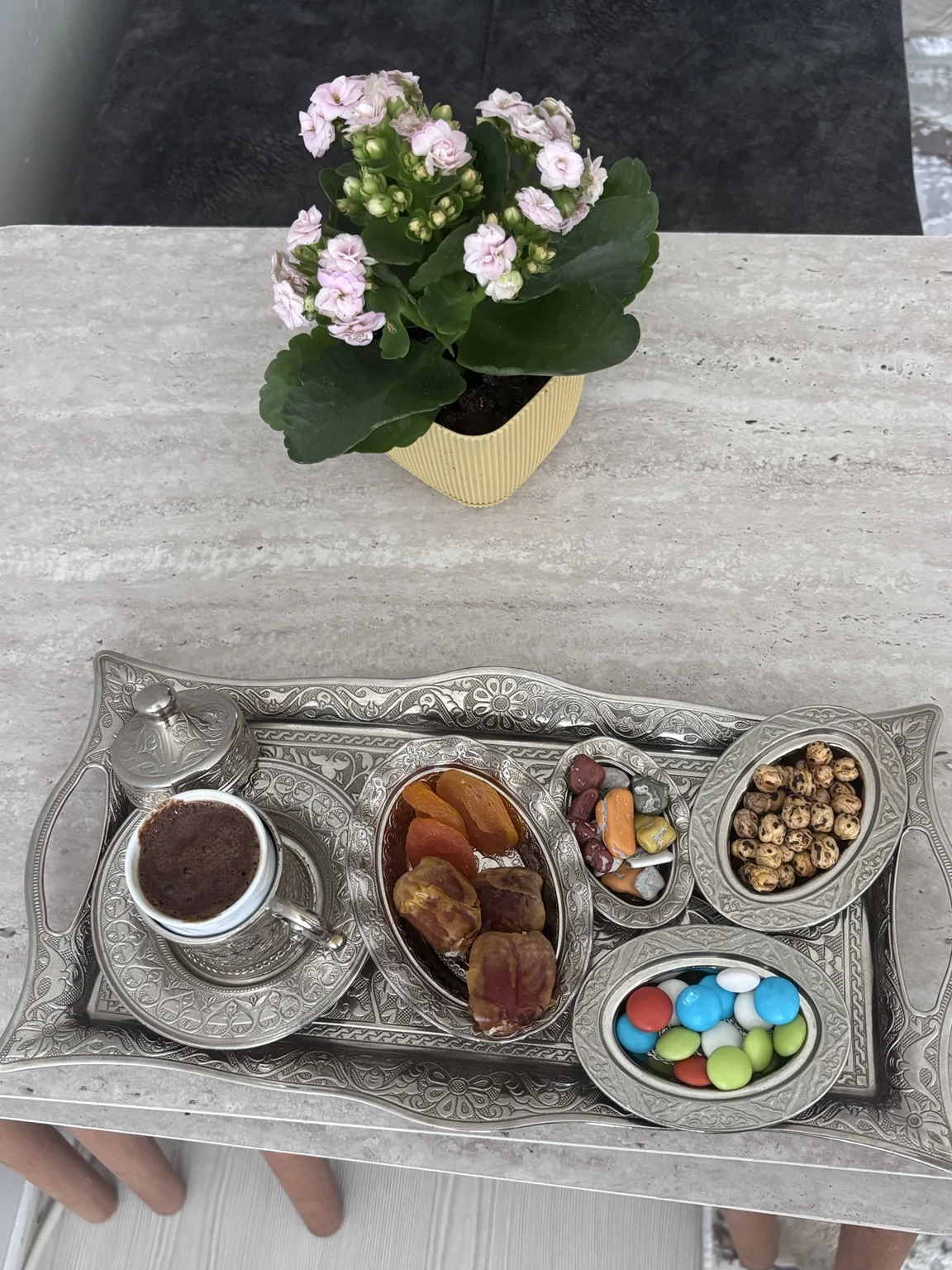 Gümüş renkli tepside sade Türk kahvesi, kuru kayısı, hurma, renkli şekerlemeler ve Tavşanlı leblebisi; arkada pembe çiçekler.